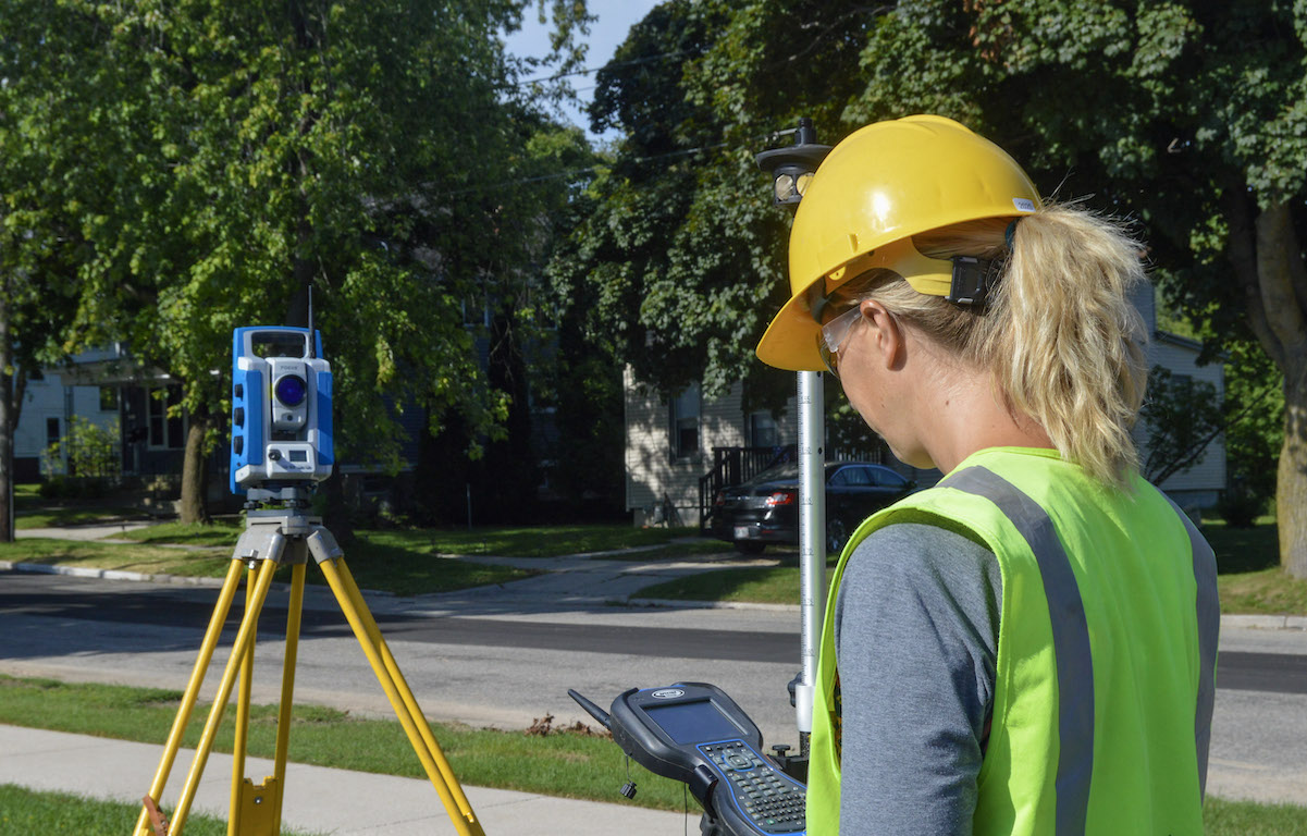 A woman wearing a safety vest and helmet works survey equipment.