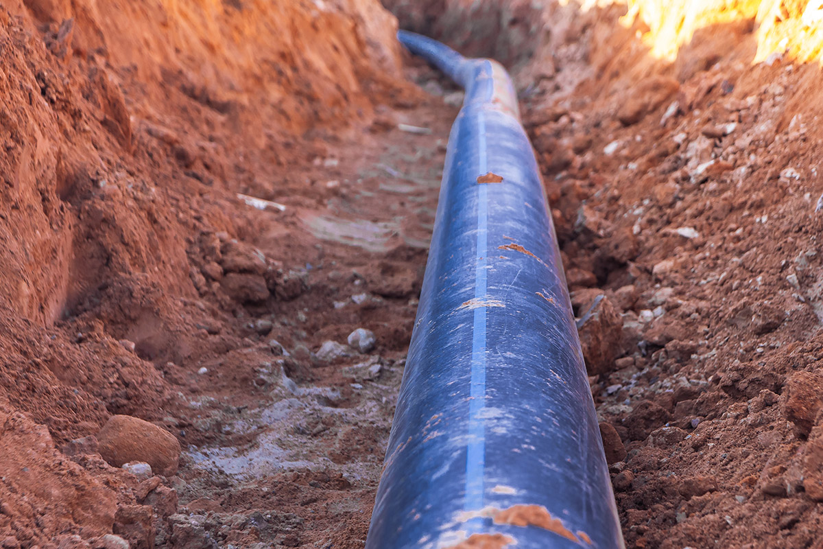 A black plastic pipe with a blue stripe lies in a dug trench to supply water to the house.